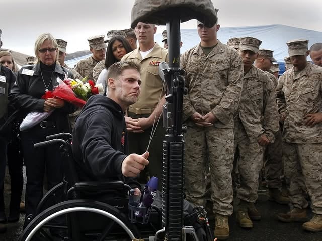 U.S. Marine LCpl Cody Elliot, with 1st Battalion, 5th Marines, holds the dog tags of LCpl Joshua B. McDaniels, a combat engineer killed during their seven-month deployment to Sangin, Afghanistan, in 2011.

1/5, along with LCpl Elliot, previously deployed to the Nawa-I-Barakzayi District of Helmand Province from May–December 2009. After 14 months back home, the battalion returned to Afghanistan, this time deploying to Sangin from March–October 2011 and relieving 3/5, another unit remembered for some of the hardest fighting and highest casualty rates of the entire war.

That same intensity carried into 1/5’s deployment. 
By 2011, Sangin had become infamous among British and American forces. It was a major Taliban stronghold, strategically important for insurgent movement routes, opium production, and its location along the Helmand River valley.

The area was filled with IEDs, canal systems, mud compounds, and dense green-zone vegetation that made ambushes common. Many who served there described Sangin as a place where even movement itself was dangerous. Patrols could take hours to move only a few hundred meters as engineers and infantry searched continuously for IEDs. Most casualties came not during major firefights, but from hidden IEDs encountered on routine patrols.

On June 12, LCpl Elliot and his platoon were moving to support sister platoons in heavy contact. After traveling only a few hundred meters outside the wire, combat engineer LCpl McDaniels triggered an IED and was mortally wounded.

After seeing the explosion, LCpl Elliot moved to reach McDaniels and render first aid. Seconds later, he triggered a secondary IED, losing a leg, a finger, and suffering severe facial injuries. Both Marines were 21 yrs old.

McDaniels was among 17 Marines killed in Sangin with 1/5 and its attached units during the deployment. More than 160 members of the battalion’s force of roughly 1,000 Marines and sailors earned Purple Hearts for combat wounds.

On June 13, 2022 (slide 7), Elliot returned to Pendleton with other vets and family of 1/5 who fought in Sangin, to hike up First Sgts Hill to lay mementos at crosses honoring members of their unit who died in combat.